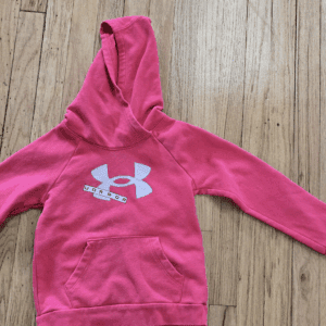 Pink Under Armour hoodie laid flat on wooden floor.