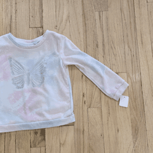 White long-sleeve children's shirt with pastel butterfly design on wooden floor.